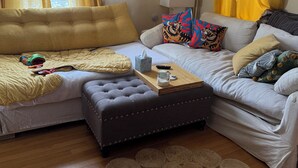 Una sala de estar con sofá, reposapiés y una mesa de centro. Hay cortinas, una lámpara y decoración en la pared.