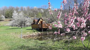 Une cabane rustique en bois, avec un toit de chaume, située au bord d’un petit étang, entourée d’arbres en fleurs et sous un ciel bleu clair.