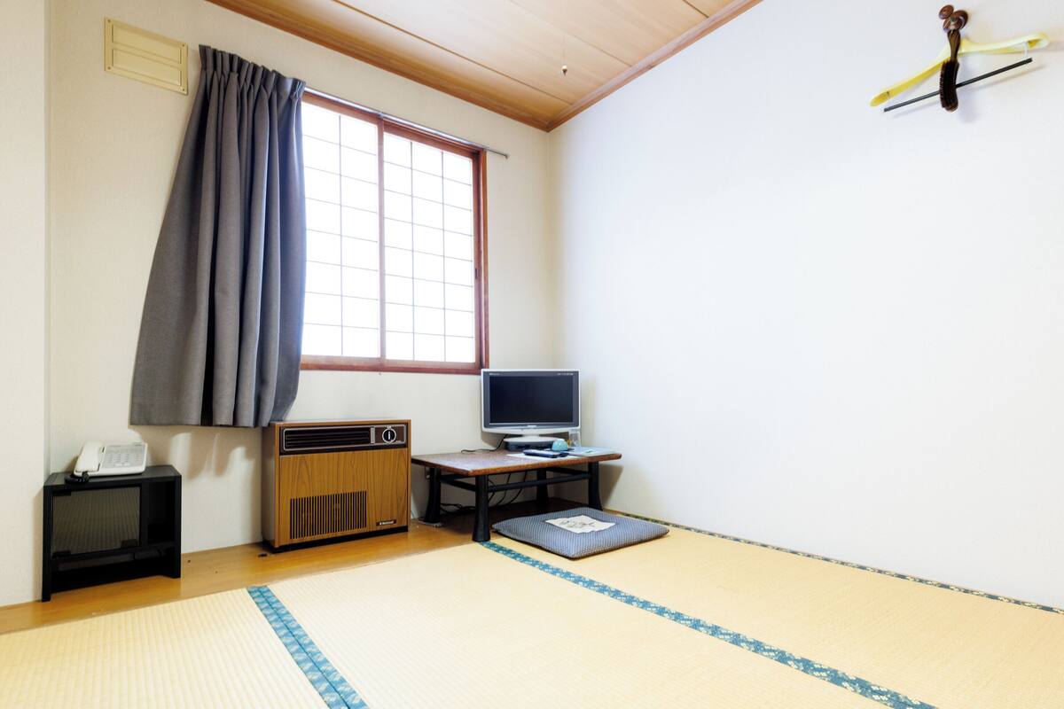 Ein traditioneller japanischer Raum mit Tatami-Boden, einem Fernseher, einer Holzschrank und einem Fenster mit Vorhängen.