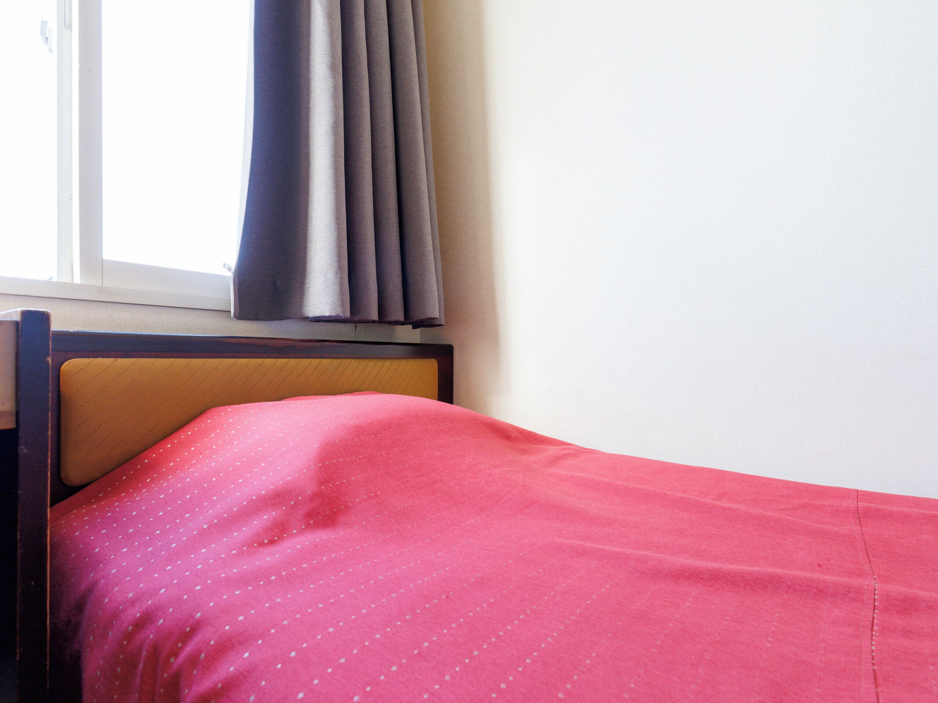 A bed with a red polka-dotted bedspread, a wooden headboard, and a window with grey curtains.