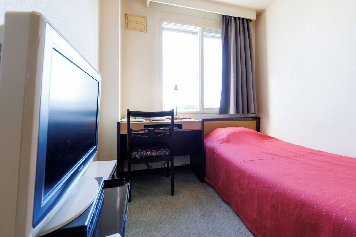 Ein kleines Hotelzimmer mit Bett, Schreibtisch mit Stuhl, Fernseher und einem Fenster mit Vorhängen.