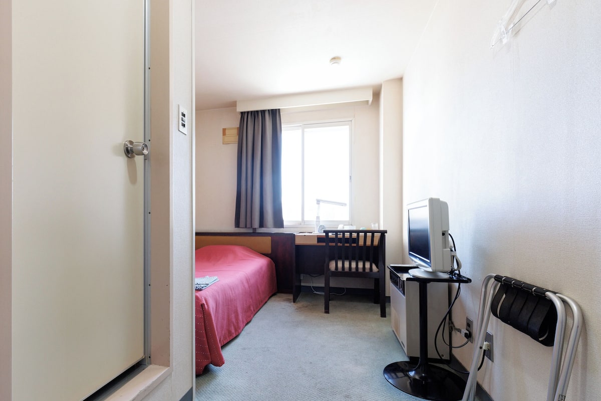 Ein Hotelzimmer mit einem Bett, einem Schreibtisch mit Computer, einem Stuhl und einem Fenster mit Vorhängen.