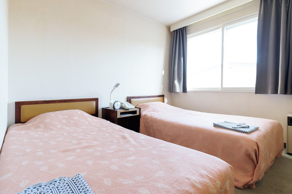 Ein Hotelzimmer mit zwei Betten, einem Nachttisch mit Lampe, einem Telefon und einem Fenster mit Vorhängen.