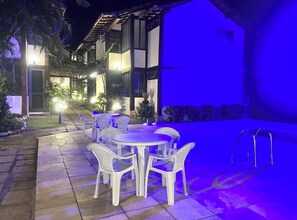 An outdoor patio with white plastic chairs and tables, a stone pathway, and a building with multiple windows and a blue-lit exterior. - House in Búzios (Geribá) a few meters from the beach. (Armação dos Búzios)