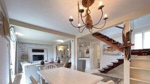 A spacious living area with a white dining table, striped chairs, and a chandelier. A staircase with wooden railings leads to a lofted space. - House in Búzios (Geribá) a few meters from the beach. (Armação dos Búzios)