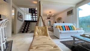 A spacious living room with a white sofa, a long beige sofa, and a glass coffee table. There is a white chair with a woven backrest and a decorative wall hanging. - House in Búzios (Geribá) a few meters from the beach. (Armação dos Búzios)