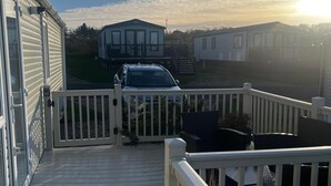 A deck with a white railing, a vehicle parked in front, and a row of mobile homes in the background.