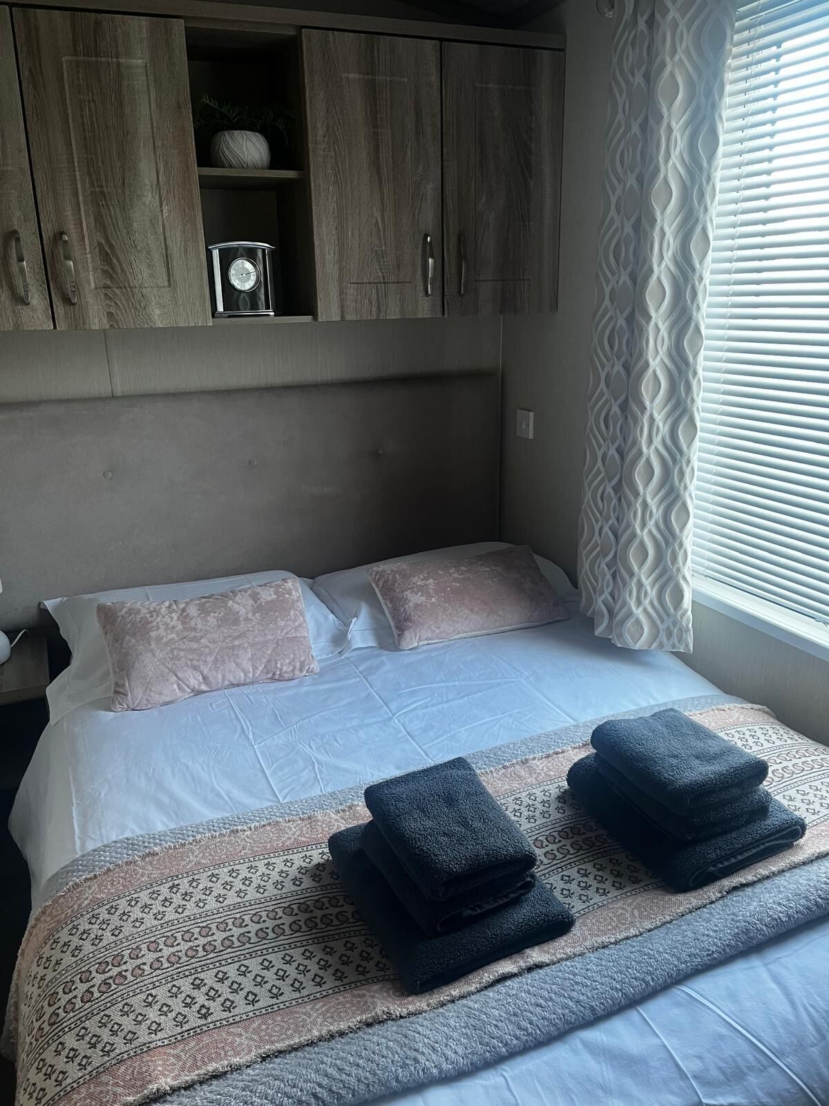 Ein Hotelzimmer mit Bett, zwei Kissen, Nachttisch, Lampe, Fenster mit Jalousien und einem Kleiderschrank.