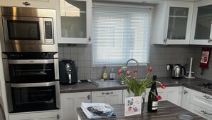 A modern kitchen with white cabinetry, a built-in oven, a microwave, and a coffee maker. There is a window with blinds, a sink, and a small island with a bottle of wine and tulips.