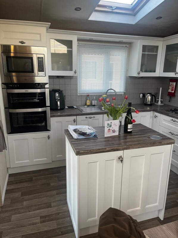 A modern kitchen with white cabinetry, a built-in oven, a microwave, and a coffee maker. There is a window with blinds, a sink, and a small island with a bottle of wine and tulips.