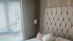 A neatly made bed with a tufted headboard, a bedside lamp, and a window with blinds.