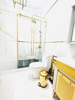Un baño moderno con ducha de vidrio, inodoro blanco y un cubo de basura dorado.