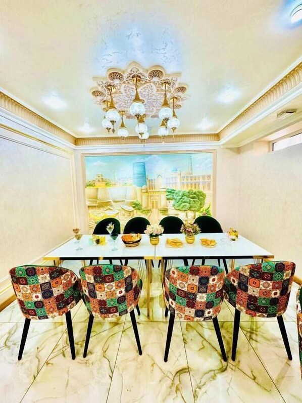 A dining area with a white table, colorful chairs, a chandelier, and a cityscape painting on the wall. - Turon Khiva (Khiva)