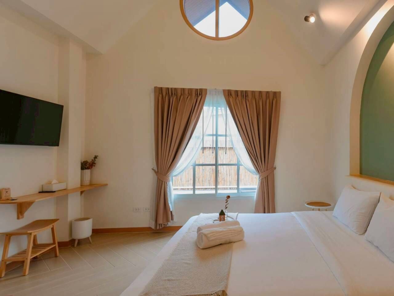 A bedroom with a large bed, a TV mounted on the wall, a window with curtains, and a small table with a plant.