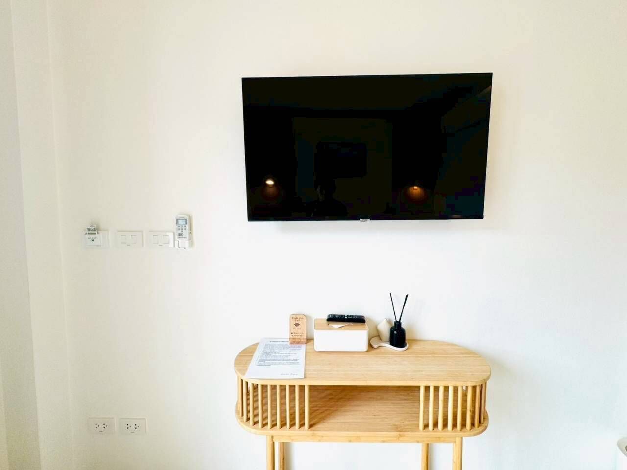 A room with a mounted TV, a wooden console table with a white box and a black device, and a small wooden block with text.