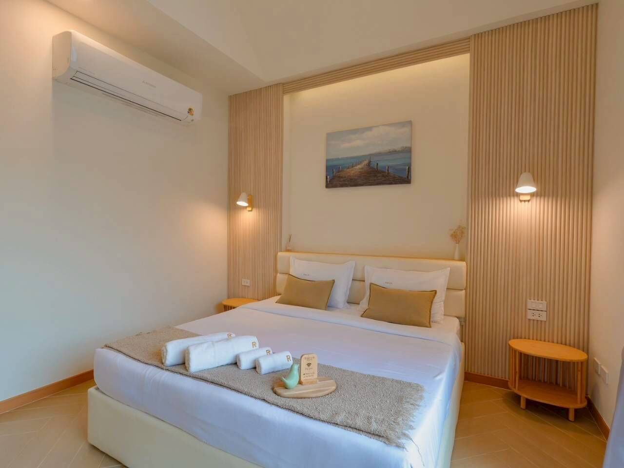 A hotel room with a bed, a bedside table, a wall-mounted air conditioner, a painting, and a lamp.