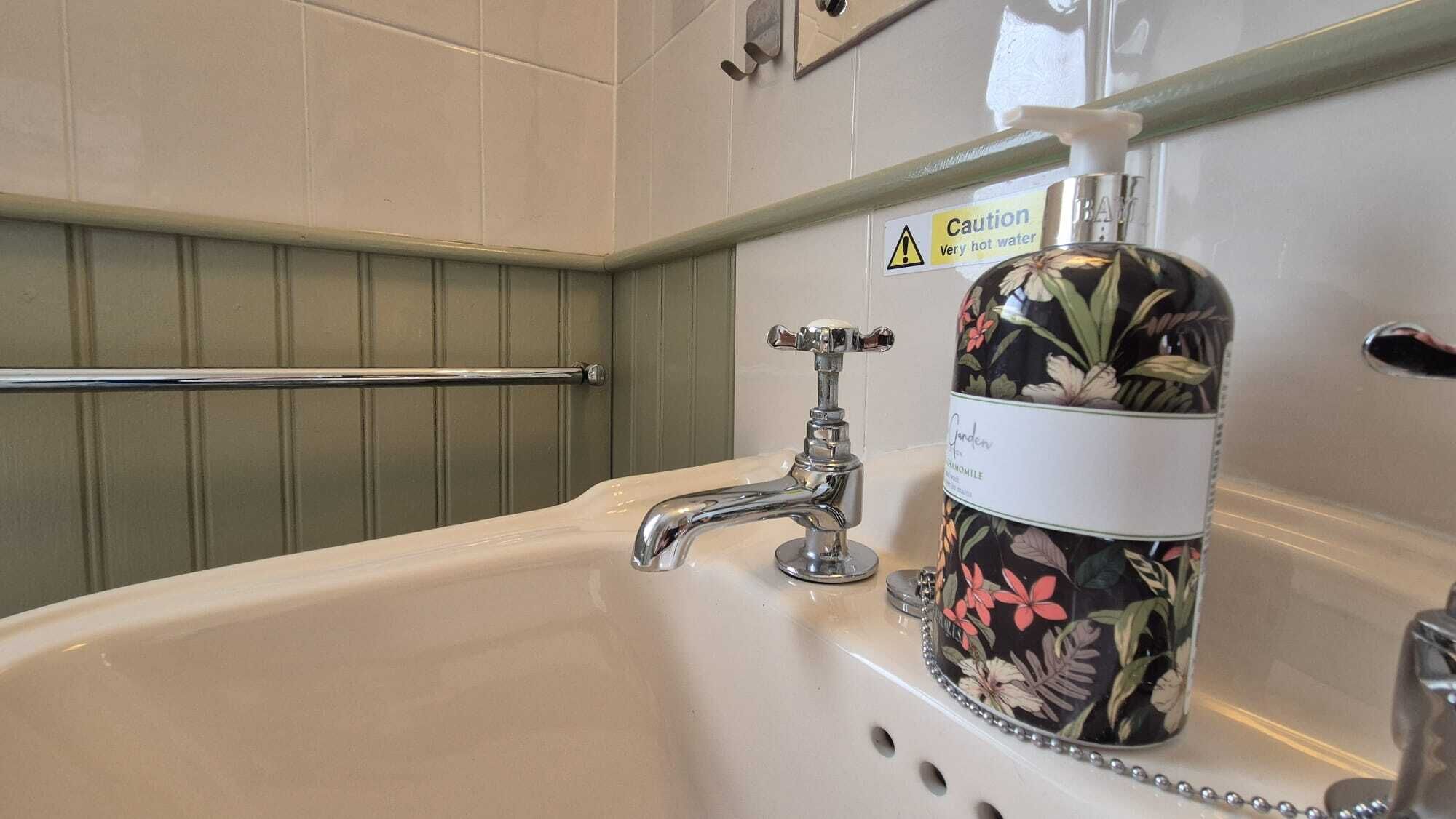 Un bagno con un lavandino bianco, un rubinetto cromato, un dispenser di sapone per le mani e un cartello di avvertimento per l'acqua calda.