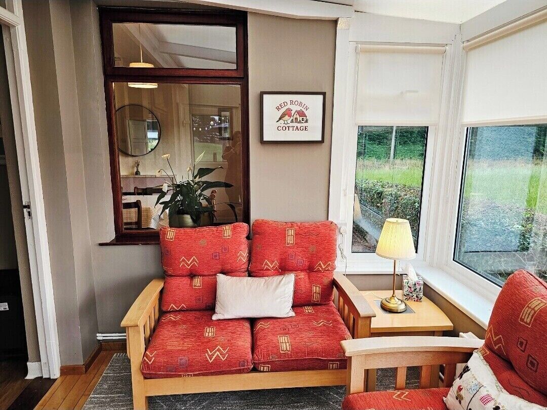 Une pièce chaleureuse avec un canapé rouge à motifs, une chaise en bois, une petite table avec une lampe et une fenêtre donnant sur un paysage verdoyant.