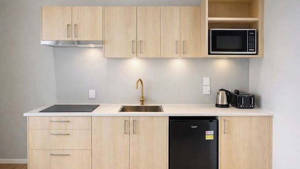 A modern kitchen with light wood cabinets, a stainless steel sink, a black dishwasher, a microwave, a kettle, and a toaster. - Rayya Suites, Paihia (Paihia)