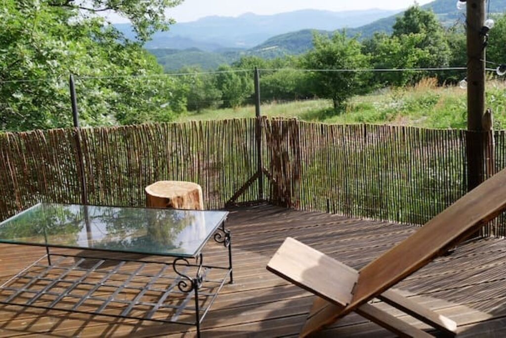Una terraza de madera con una mesa de vidrio y una silla de madera, con vistas a una cerca de bambú y un paisaje montañoso.