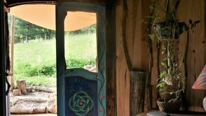 Une porte en bois rustique, ornée d’un motif décoratif, qui donne sur un espace extérieur comprenant un champ d’herbe et un banc en bois sur lequel est posée une plante en pot.