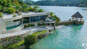 A modern waterfront property with a multi-level structure, a pool, and a thatched-roof gazebo.