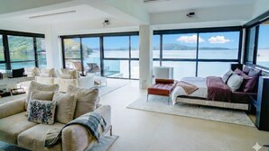 A spacious living area with a large bed, a sofa, and a coffee table. There are large windows offering a view of the water.