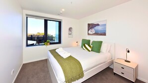 Un dormitorio con un ventanal grande, una cama con almohadas verdes, una mesita de noche con lámpara y un cuadro en la pared.