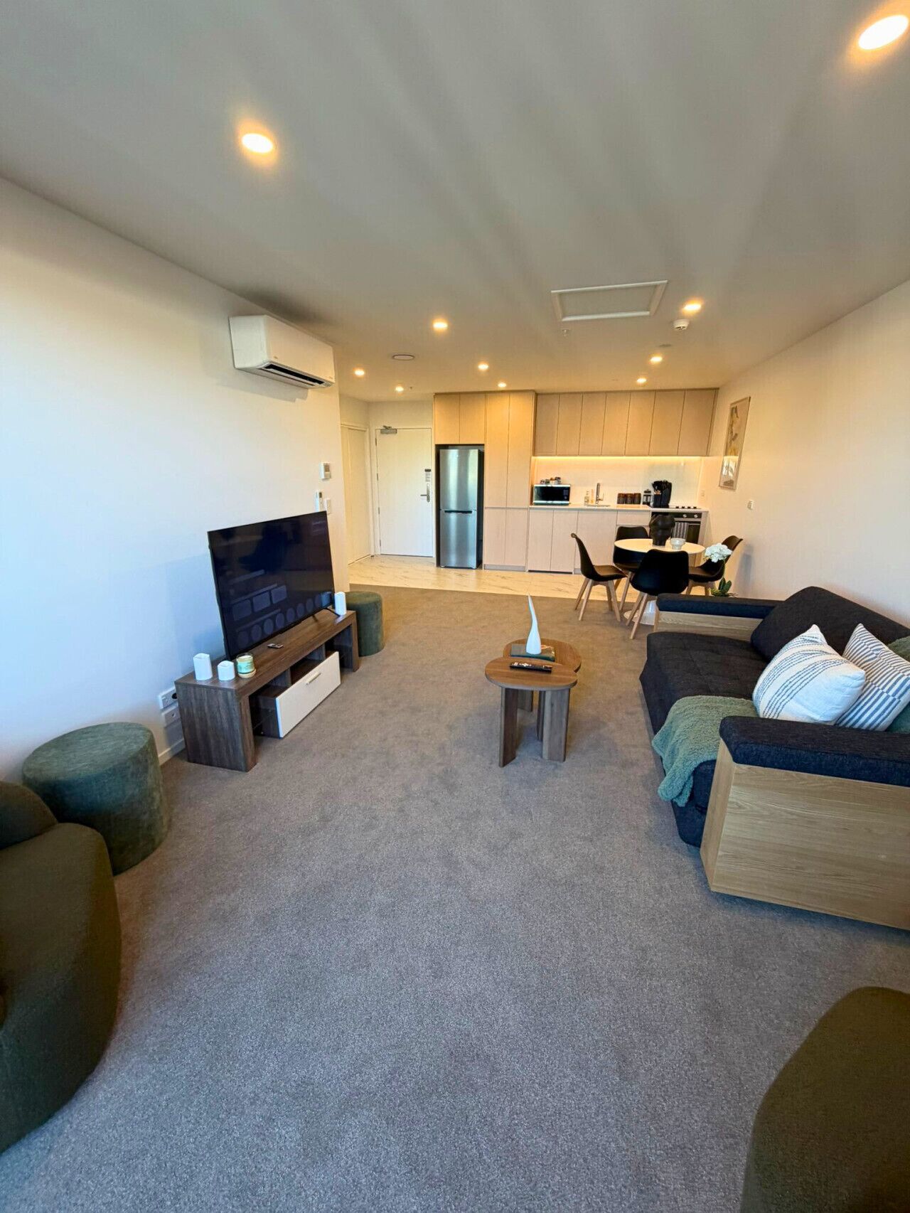 Una sala moderna con televisor de pantalla plana, un sofá y una mesa de centro. Se ve una cocina al fondo con refrigerador y microondas.