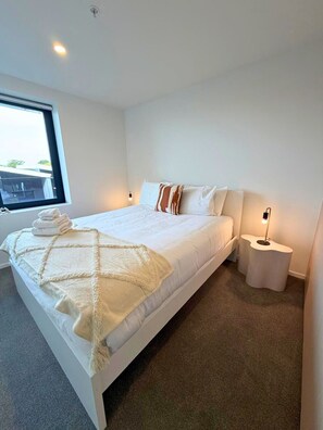 Una habitación de hotel moderna con una cama grande, una mesita de noche con lámpara y un ventanal con vistas al exterior.