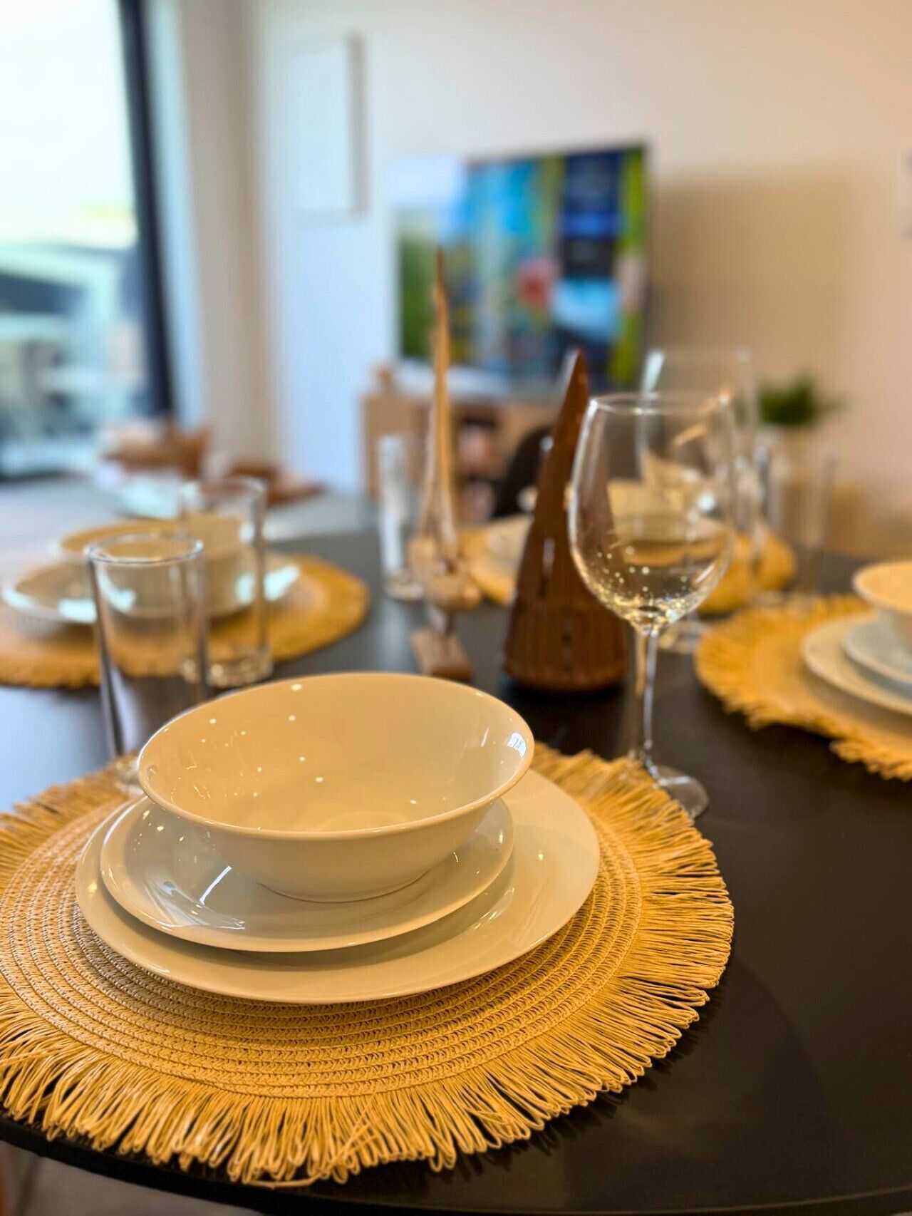 A dining table set with white plates, bowls, and wine glasses, with a woven placemat underneath.
