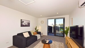 A modern living room with a dark sofa, a coffee table, a flat-screen TV, and a large sliding glass door leading to a balcony. - Fresh Build City Retreat in Christchurch CBD (Christchurch)