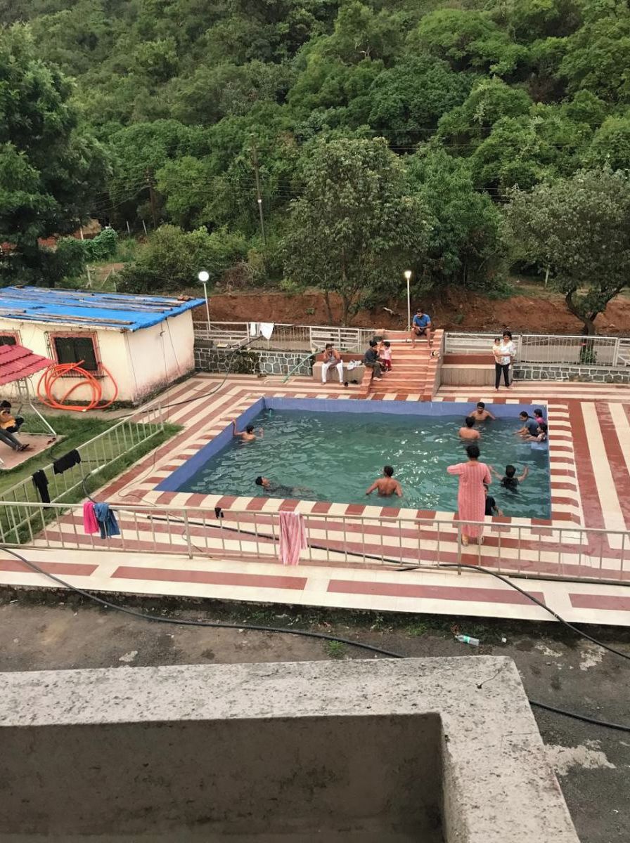 A swimming pool area with people swimming and sitting around. There is a small building with a blue roof and a hose coiled beside it.