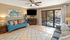 A living room with a blue sofa, a grey sofa, a ceiling fan, a flat-screen TV, and a large window with curtains. - PCB Condo Near Pier Park + Pool + Pickleball Court | Beachwalk 10B by AvantStay (Panama City Beach)