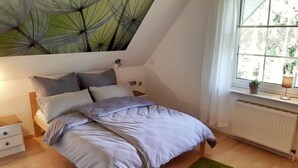 Une chambre avec un plafond en pente, un lit avec une literie grise, un tapis vert, une table de chevet et une fenêtre donnant sur un paysage verdoyant.