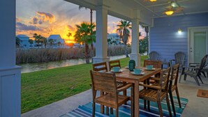 Un patio techado con mesa y sillas de madera, con vista a una zona verde y un cuerpo de agua con casas al fondo.