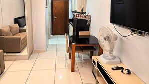 A modern living room with a sofa, a wooden chair, a TV mounted on the wall, and a standing fan. - Budget Studio Condo with Resort-Style Pool - Laguna Beach Resort 1, Jomtien (Muang Pattaya)