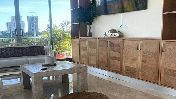 A modern living room with a large TV, wooden cabinets, and a city view. - Luxurious 3-Bedroom Apartment in Juan Dolio with Pool Access (Juan Dolio)