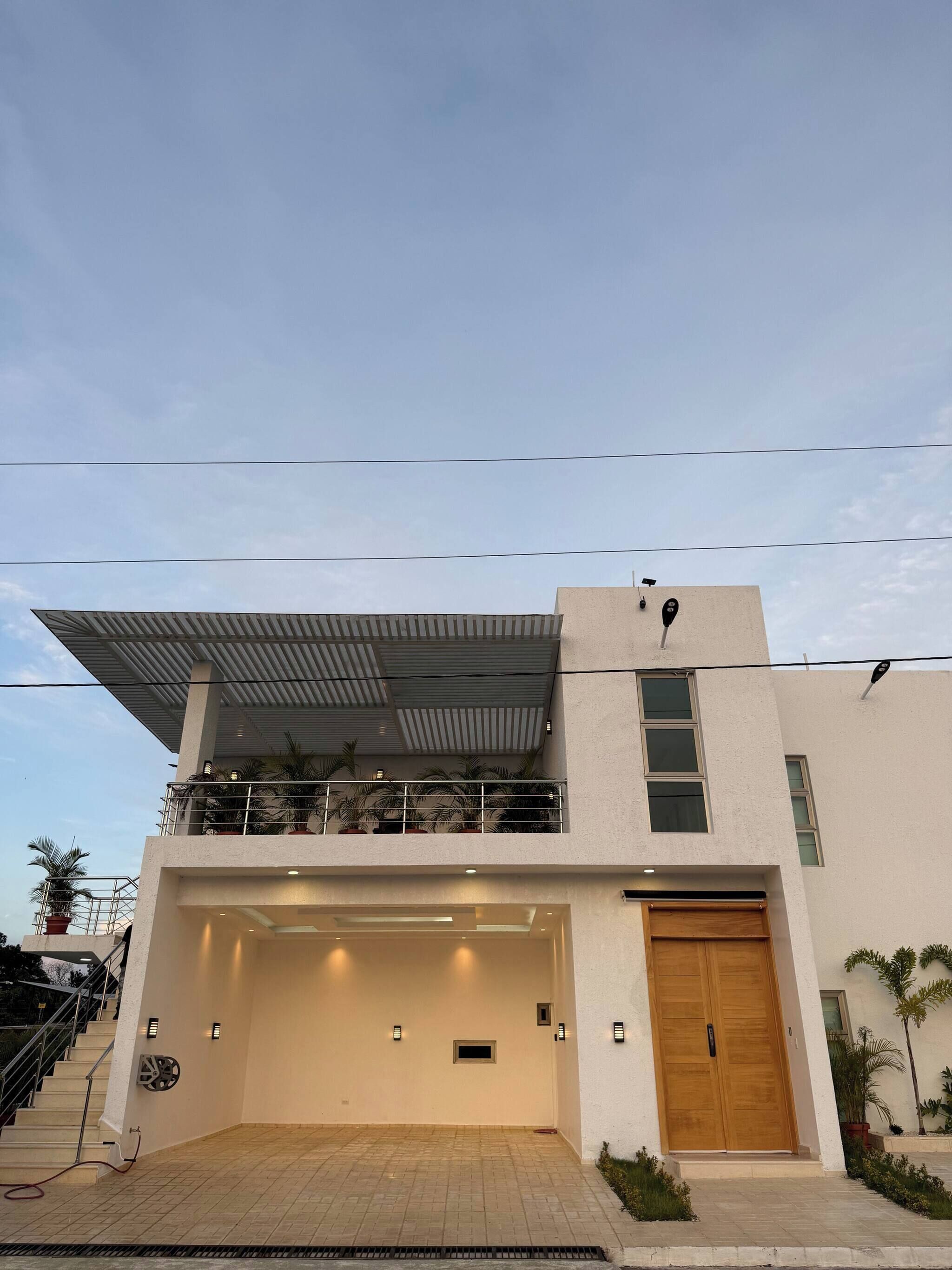 A modern two-story building with a flat roof, a covered balcony with potted plants, a garage entrance, and a wooden door.