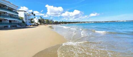 A beachfront hotel with multiple floors and balconies, a sandy beach, and clear blue skies.