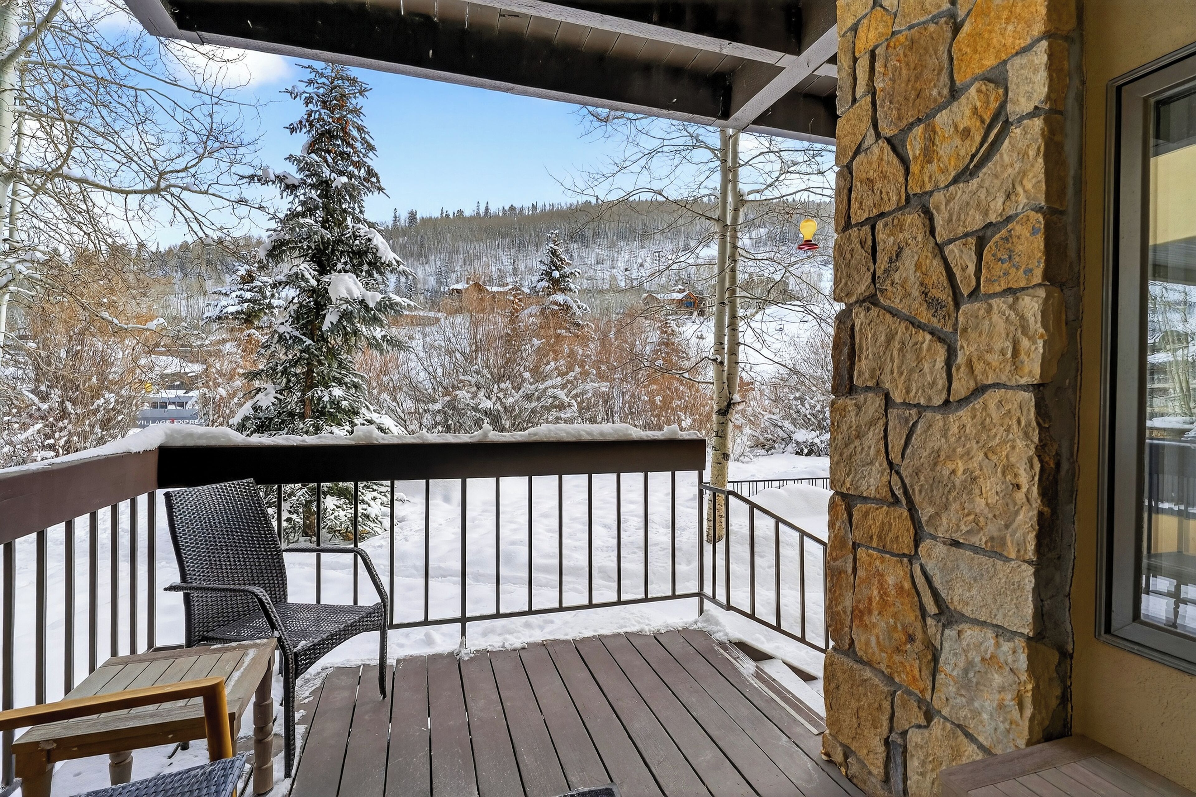 Une terrasse en bois avec un mur en pierre, une chaise et une vue sur des arbres et des bâtiments recouverts de neige.