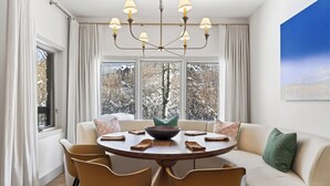 Uma sala de jantar com uma mesa redonda, cadeiras, um candelabro e um sofá com almofadas.