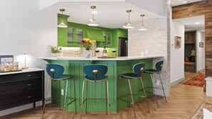 Uma cozinha moderna com armários verdes, uma ilha central com banquetas, iluminação pendente e vista para a área de estar adjacente.