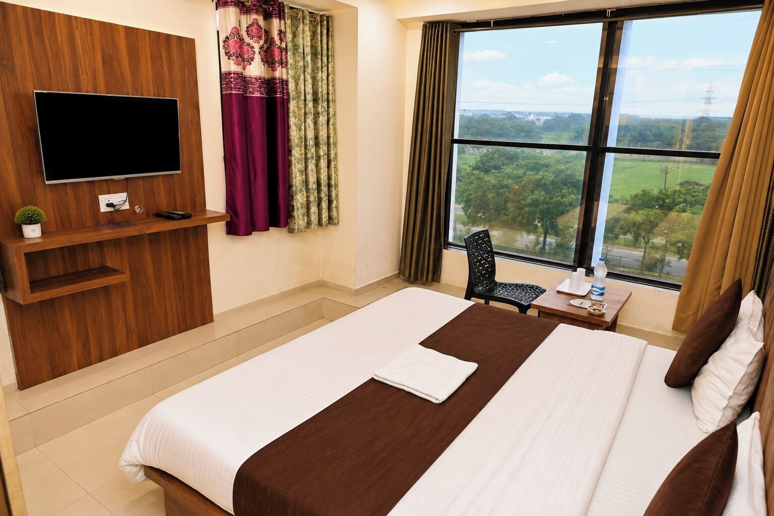 A hotel room with a large bed, a flat-screen TV mounted on the wall, a small table with a chair, and a window with a view of greenery.