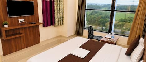 Une chambre d’hôtel dotée d’un grand lit, d’un téléviseur à écran plat fixé au mur, d’une petite table avec une chaise et d’une fenêtre donnant sur un paysage verdoyant.
