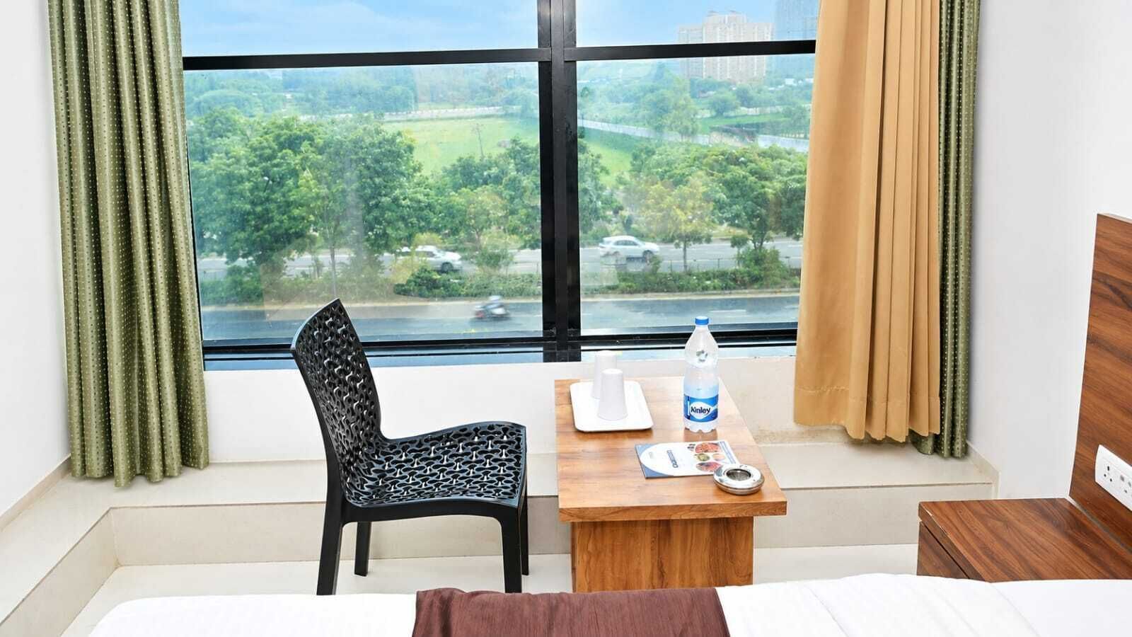 A hotel room with a bed, a wooden table with a bottle and a cup, a chair, and a window with curtains.