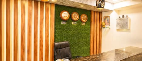 A reception desk with a wooden panel design, a green wall with clocks labeled 'INDIA,' 'U.S.A,' and 'U.K.,' and a black leather chair.