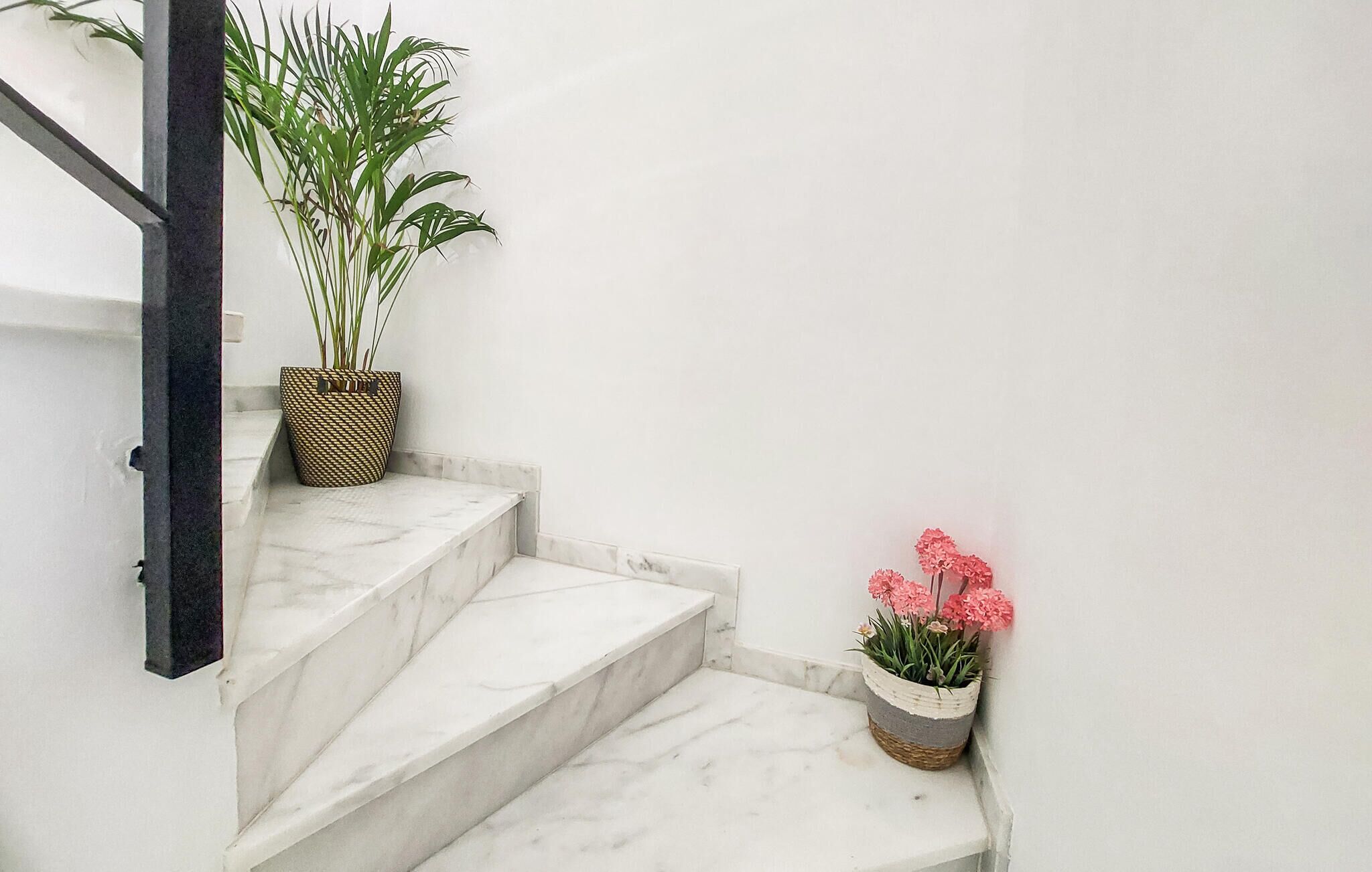 Una escalera con escalones de mármol blanco, una planta en maceta a la izquierda y una cesta con flores rosadas a la derecha.