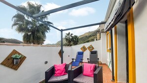 A covered outdoor seating area with two chairs and cushions, a small table with a plant, and a view of palm trees and hills. - Lovely home in Benaojan with swimming pool (Benaojan)