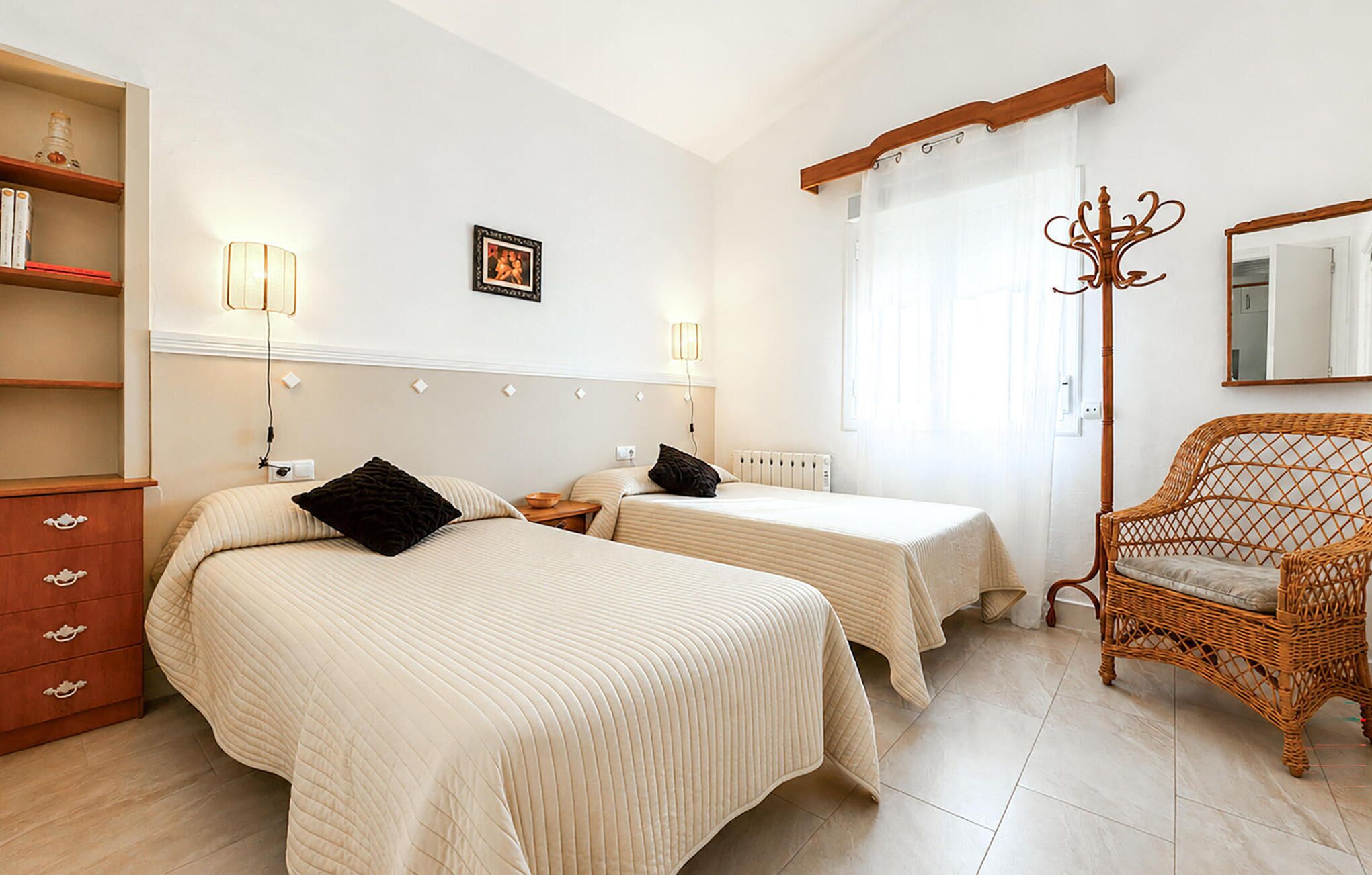A hotel room with two beds, a wooden dresser, a wicker chair, and a picture on the wall.